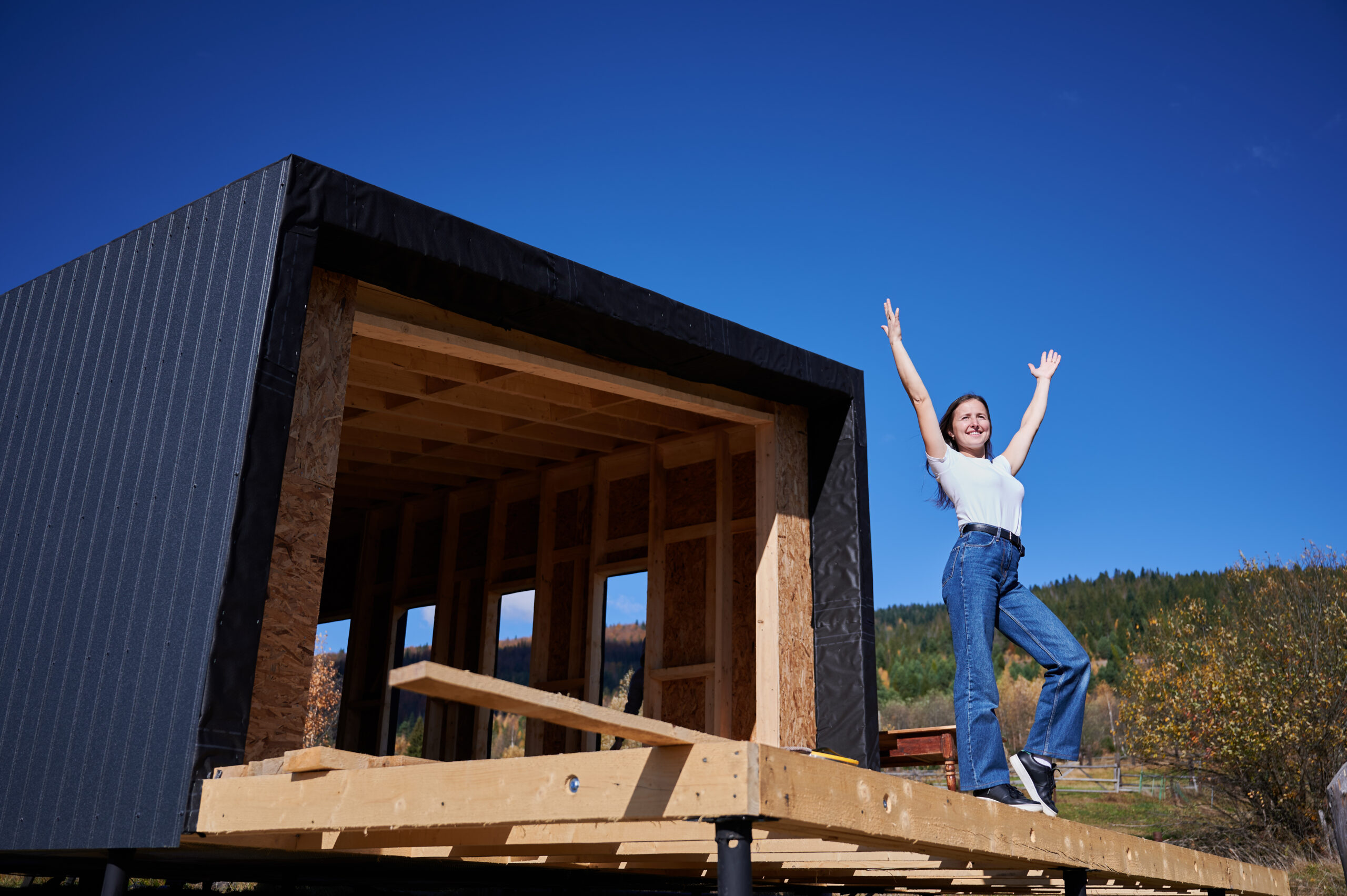 Happy woman on construction site standing on terrace at incomplete wooden frame house on sunny day with hands up.